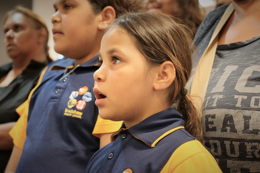 An Indigenous girl in a blue and yellow school uniform singing with a group.