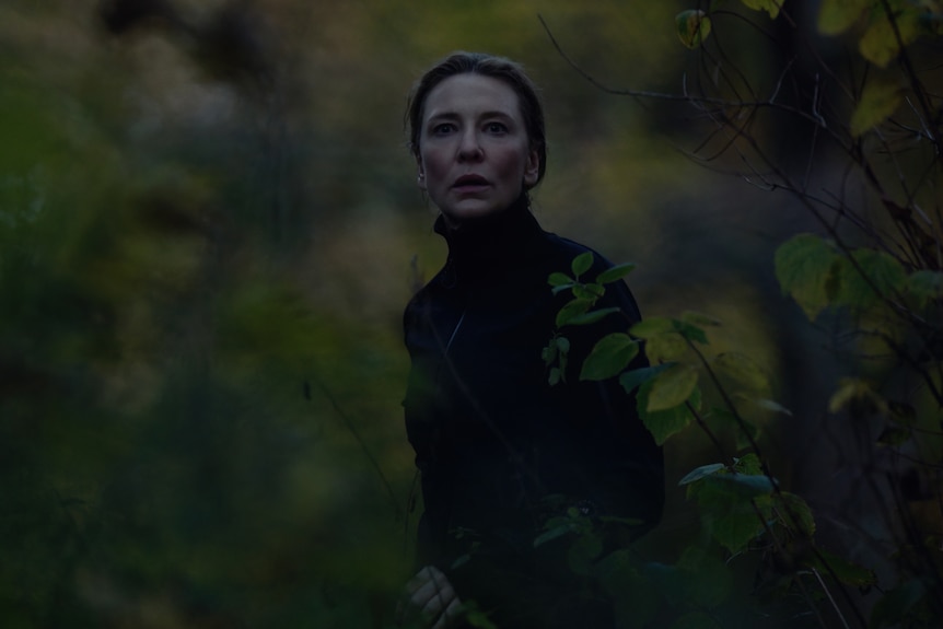 A brown-haired, 50-something woman in a turtleneck looks frightened, standing out in nature