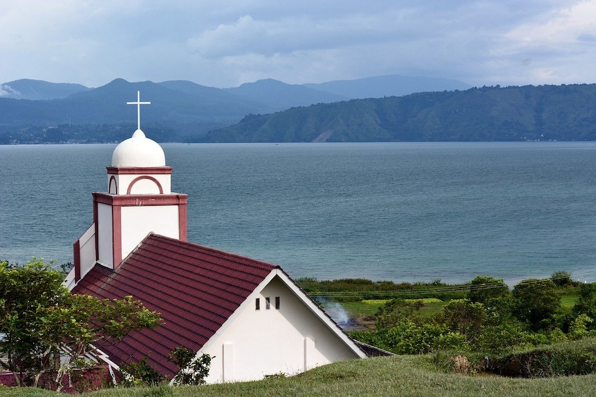 On a bright blue day, you look down at a small red-roofed Church from behind as blue waters are seen beyond it.