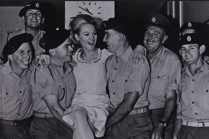Black and white photo of a woman being carried between two men in uniform, wearing berets and other troops crowding around.