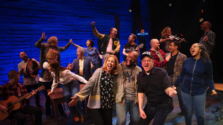 Actors in the musical Come From Away on stage, joyously singing, dancing playing musical instruments.