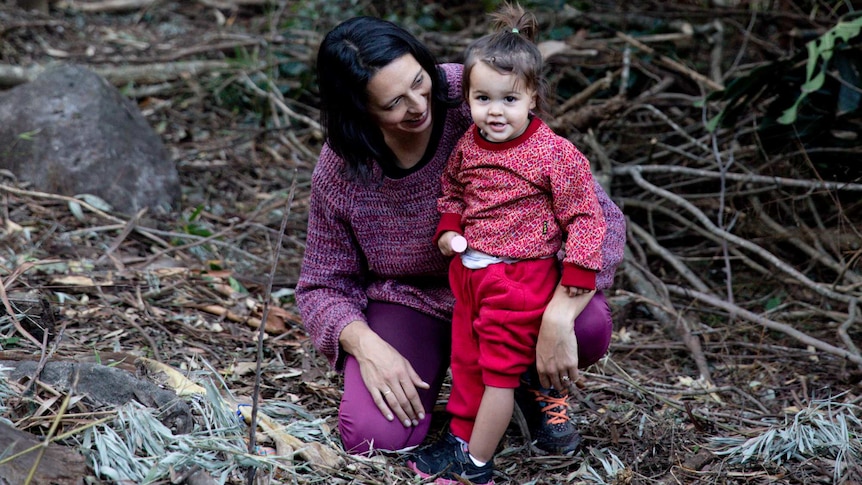 A woman with dark brown hair crouches with her young child in a bush setting.