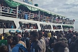 A ferry parked next to a wharf is packed with people and there is a huge crowd of people on the wharf as well.