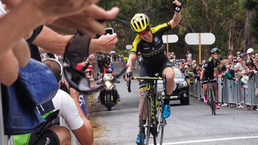 A cyclist smiles and raises her left arm in victory.
