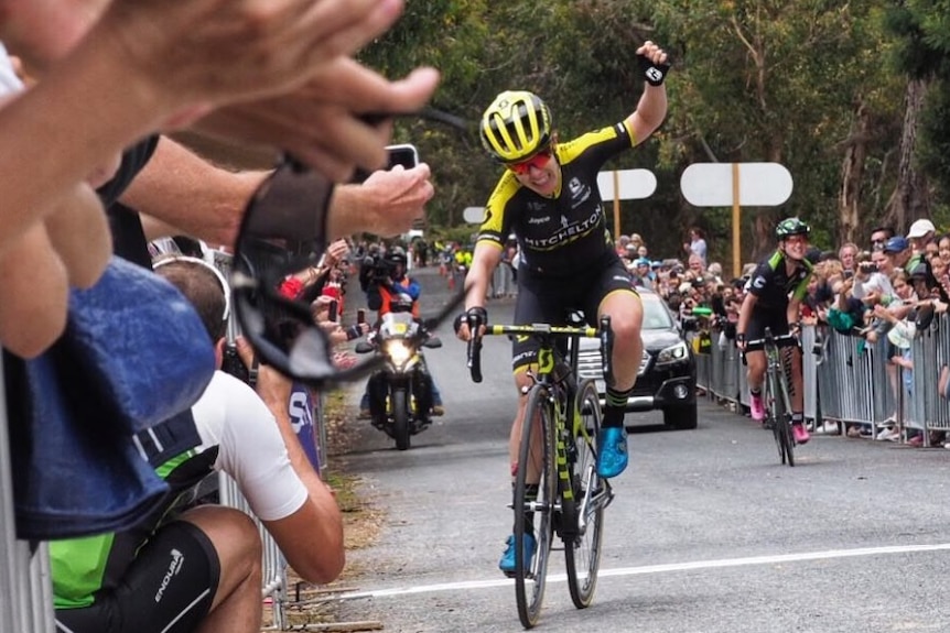 A cyclist smiles and raises her left arm in victory.