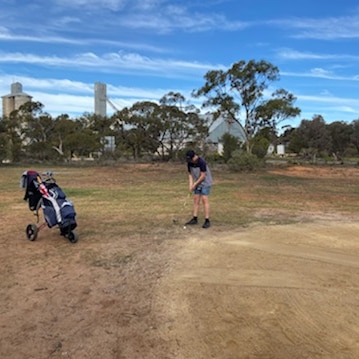 Teenage boy lines up a putt on a sand scrape at a golf course, with golf  buggy nearby and silos in background