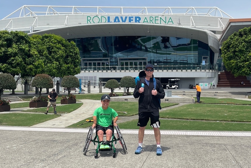 A young man with dwarfism in a wheelchair and a middle-aged man stand in front of Rod Laver Arena