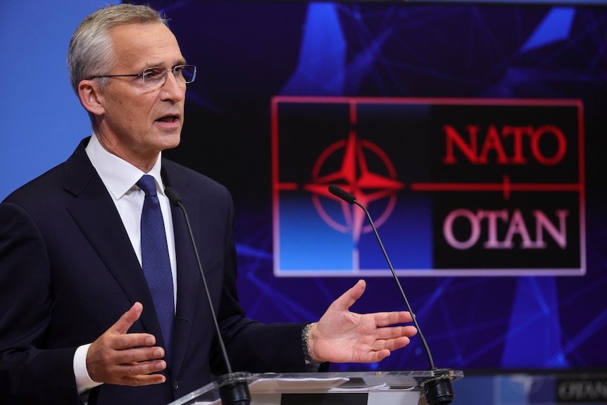 A man in a suit speaks from a lecturn near a NATO OTAN sign.