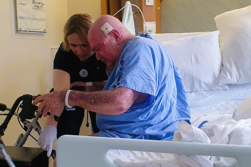 A man wearing a light blue hospital gown being assisted out of a hospital bed by a nurse wearing a navy blue uniform.