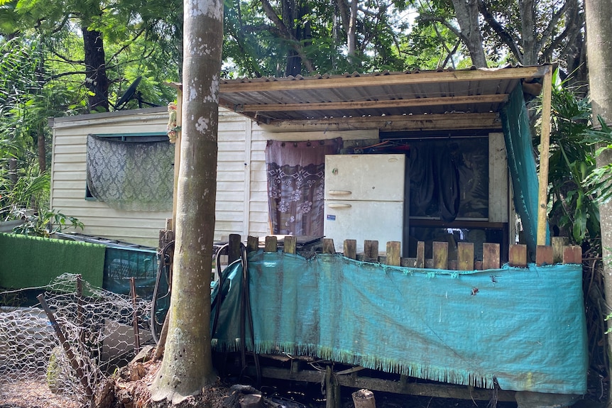 Run down white cabin with timber balcony and old white fridge in foreground. 