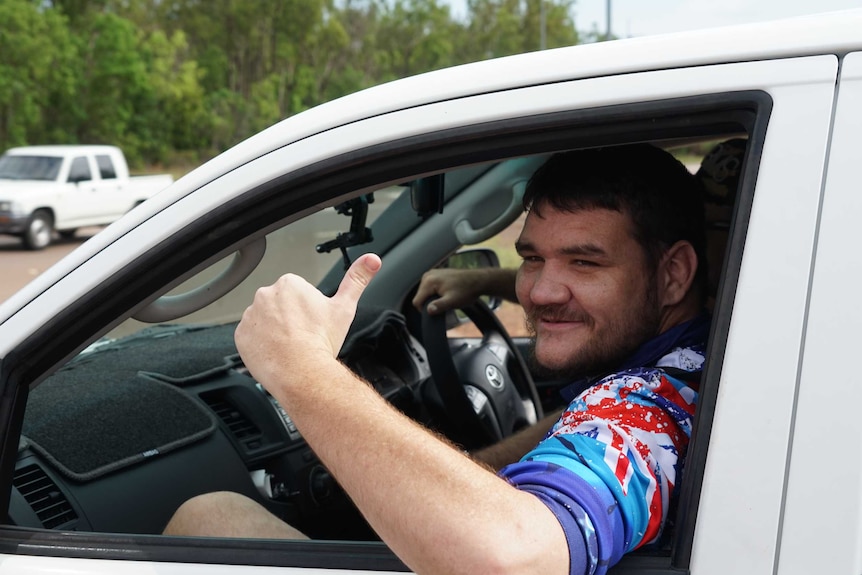 Geoffrey Watson gives a thumbs up from a white car. He looks happy and someone can be seen in the background driving the car.