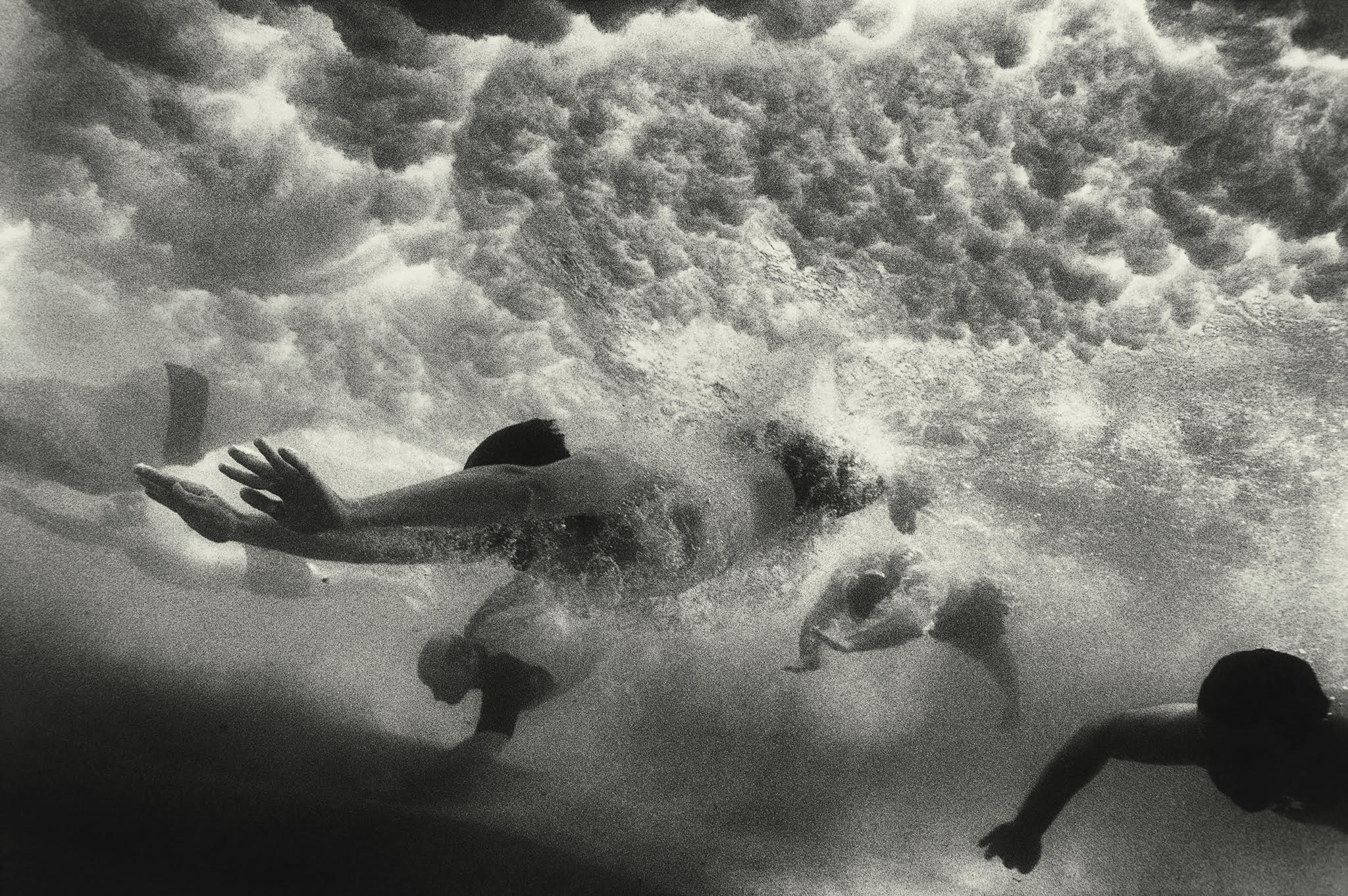 Moody black and white underwater photo by Narelle Autio of swimmers diving under a forceful wave  