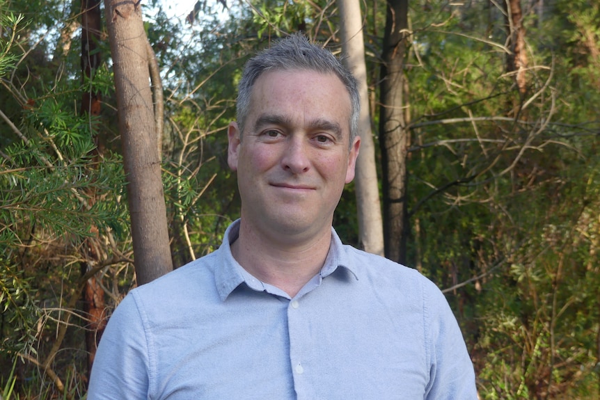 A man smiles at the camera, in front of bushland