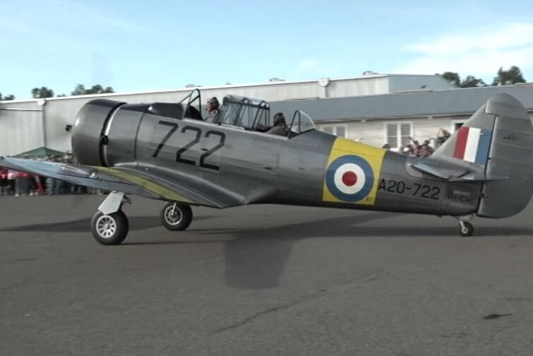 A grey Wirraway plane from World War II rolls along the tarmac, with a big shed in the background.