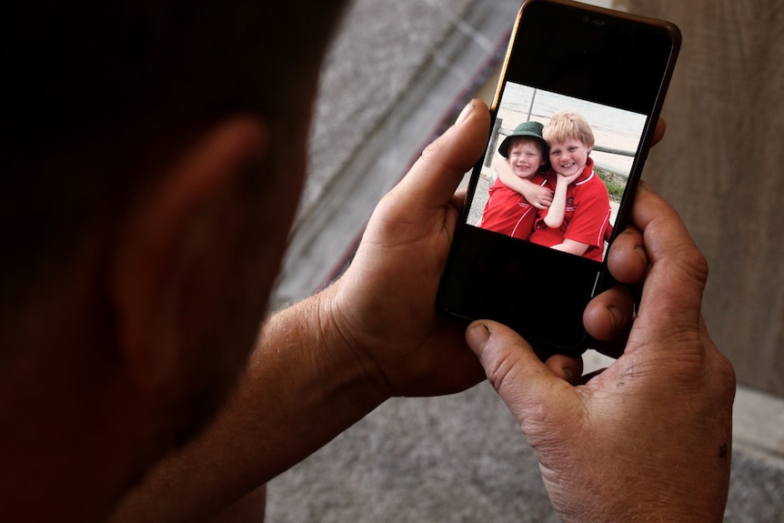 A close-up shot of a mobile phone in the hands of a man, showing a picture of a young boy and young girl wearing red shirts.