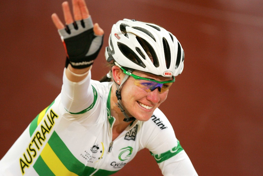 Wearing an Australian jersey, Kate Bates smiles and waves while riding her bike
