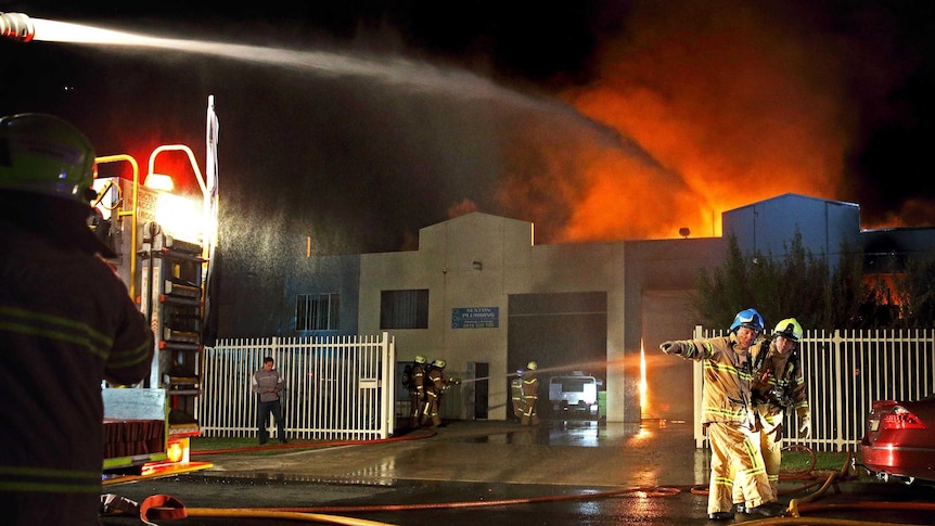 Night photo of a hose on the back of a fire engine spraying water onto fire, with crews working around the fire engine.