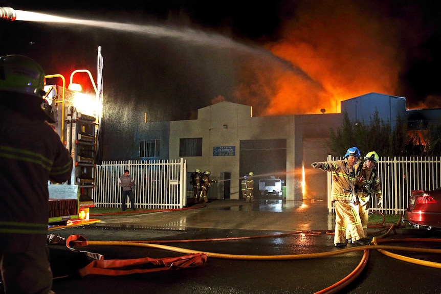 Night photo of a hose on the back of a fire engine spraying water onto fire, with crews working around the fire engine.