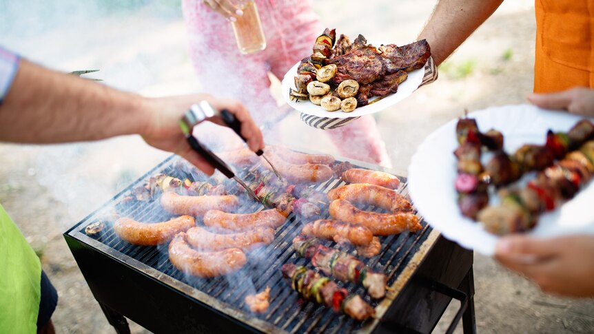 Various meats cooking on a barbecue, surrounded by people.