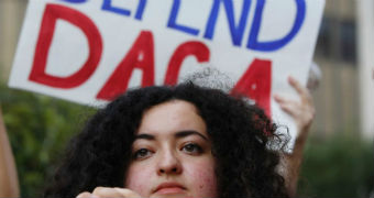A woman stands in front of a sign saying "Defend DACA".
