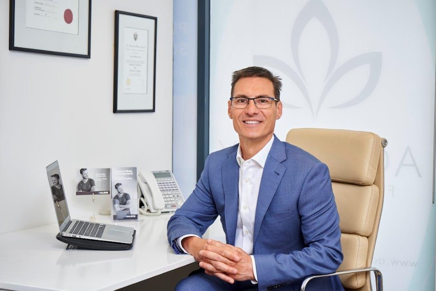 A man wearing glasses and a blue suit sits at his desk.