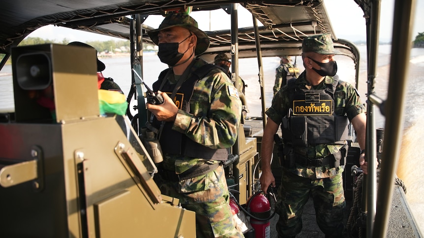 Two soldiers wearing camouflage gear, masks and vests stand aboard a patrol boat on the Mekong River