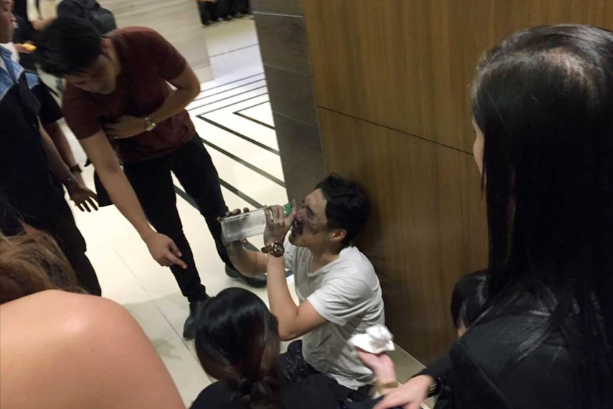 An injured man with black marks on his face drinks water while sitting on the floor.