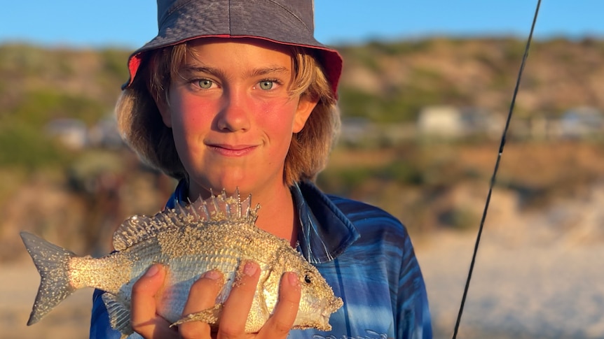 A 12 year old boy wearing a bucket hat smiles as he holds a fish with his hand.
