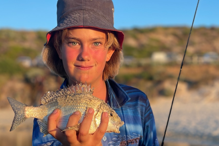 A 12 year old boy wearing a bucket hat smiles as he holds a fish with his hand.