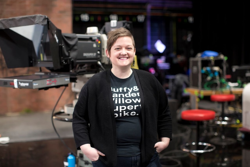 A non-binary person in a black tshirt and cardigan stands in a TV studio