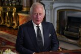 King Charles sits at a wooden table inside Buckingham Palace. He is wearing a black suit.