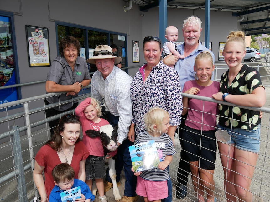 Group of farmers, supermarket workers and kids stand around cow, holding book.