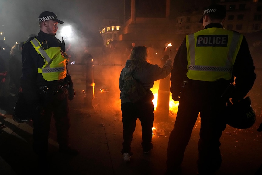 Two police oficers watch a protester, who is holding a firework and is illuminated by the glow of a nearby fire.