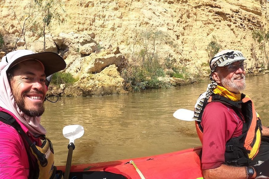 Two people sit in a kayak on the Murray River in front of a cliff. 