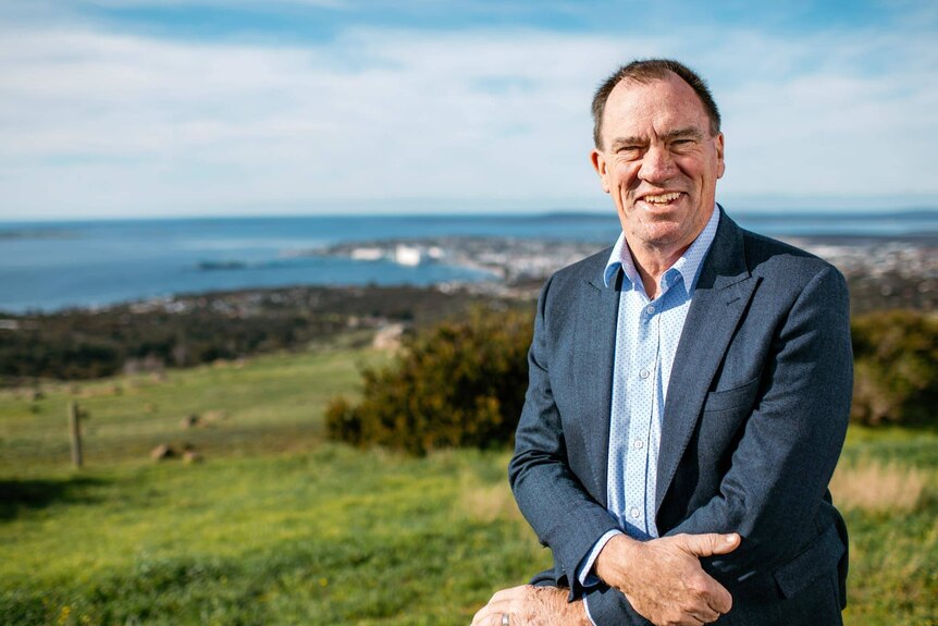 Smiling man in a a suit looking at camera from hilltop with blurred bay in background