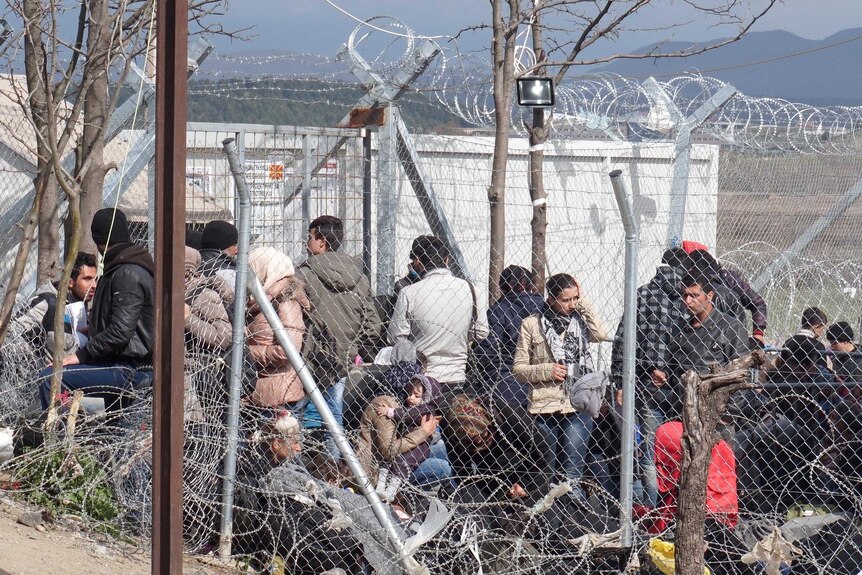 Asylum seekers sit on the ground between razor wire fences.