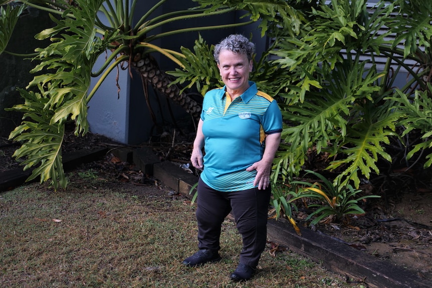 Maree, wearing a green and gold polo shirt and black pants, stands in a garden. 