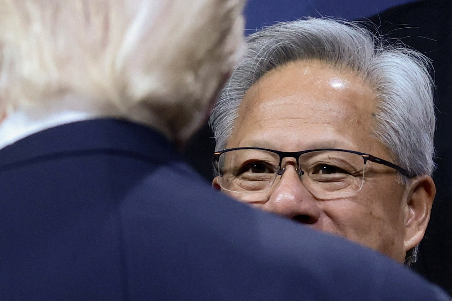 Jensen Huang, with grey hair and glasses, his face partially hidden by the shoulders and blonde hair of Donald Trump.