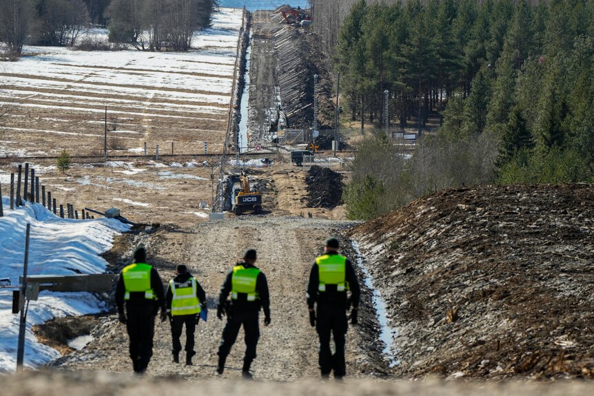Four border guards look down a dirt road as a long border fence is construced between the road and a pine forest. 