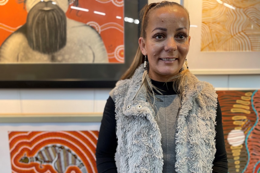 A  woman wearing a fake fur vest standing in front of Aboriginal artwork, smiles.