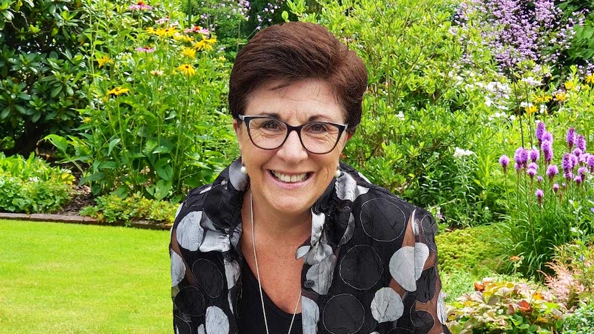 Woman wearing glasses smiles and leans forward to the camera. She is surrounded by blooming flowers