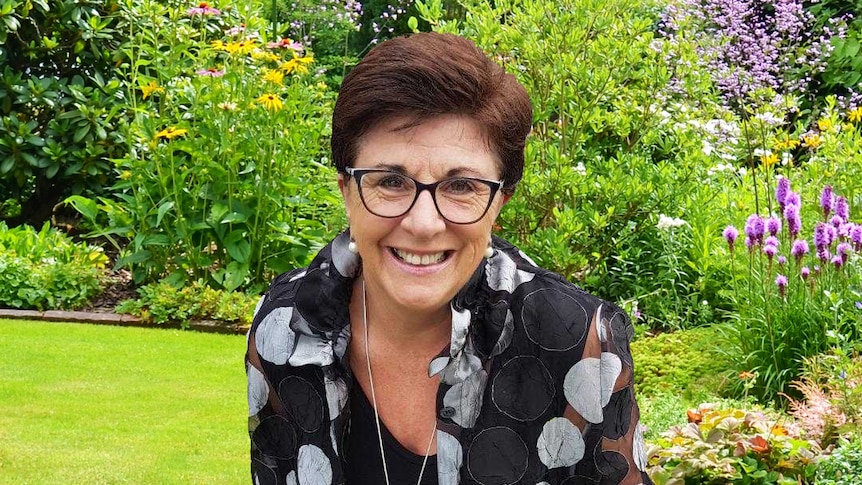 A woman sits outdoors, smiling in a beautiful garden full of flowers.