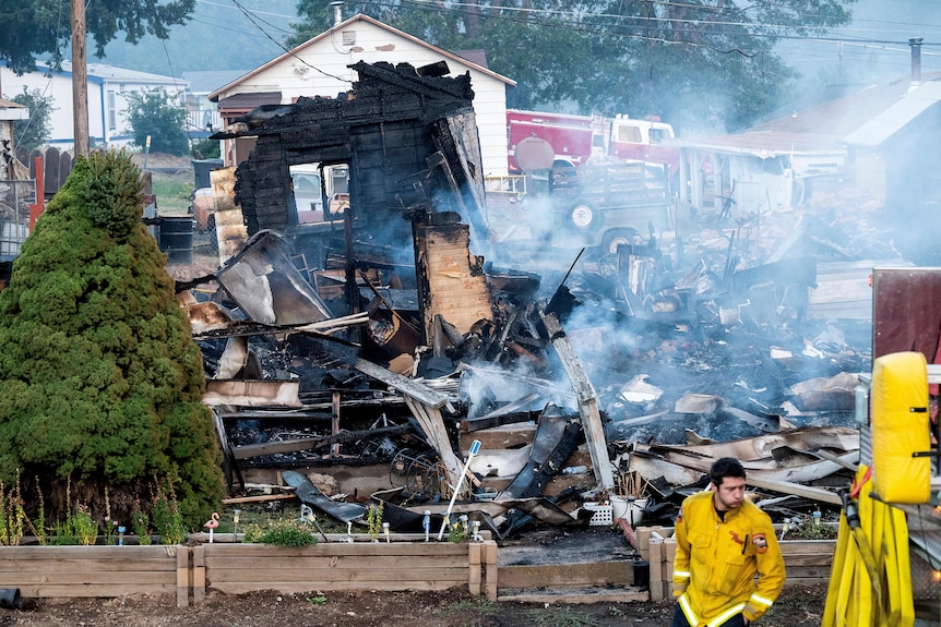A house has been gutted by fire. In the foreground is a green tree and a firefighter in a yellow uniform. 