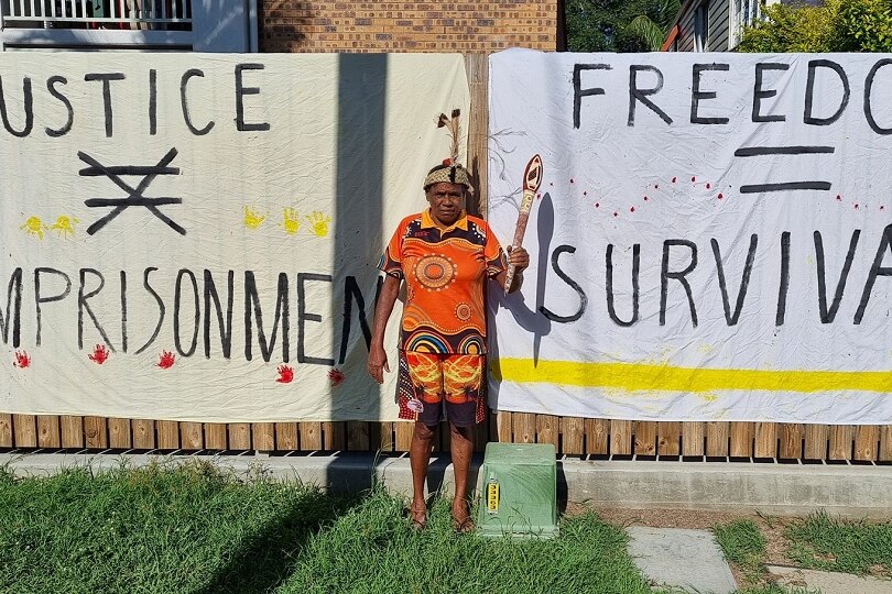 An Indigenous woman standing in front of two banners 'justice x imprisonment', holding a torch and wearing a colourful outfit.