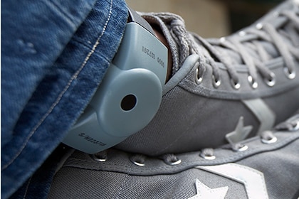 A close-up shot of an electronic tracking bracelet on a person's ankle between grey shoes and jeans.