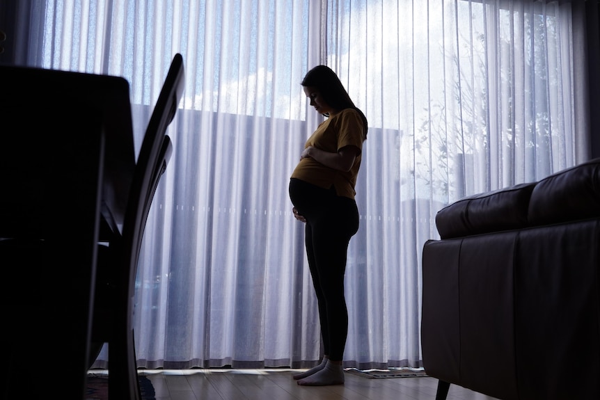Silhouette of a heavily pregnant woman standing in a lounge room against a sheer curtain