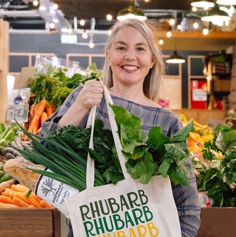 A blonde woman, in the middle of a market, holding a shopping bag filled with vegetables, smiling to the camera