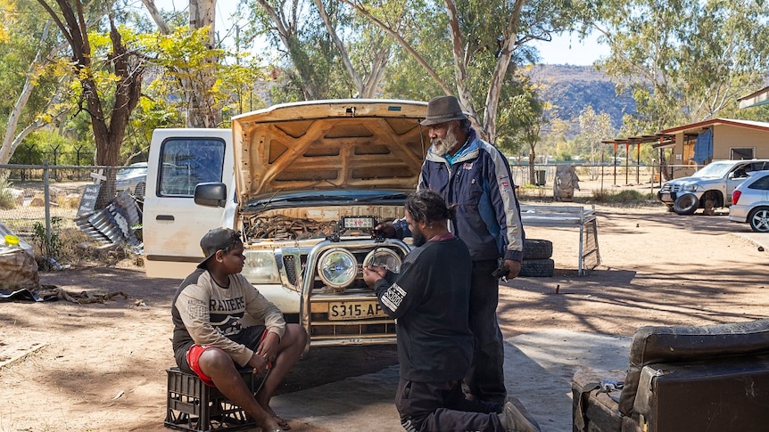 Two men in front of a car with the bonnet raised show a teenage boy how to fix a car