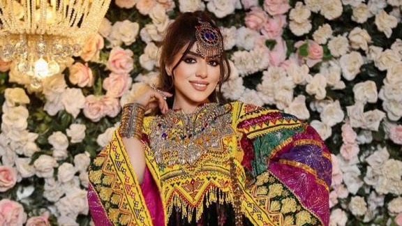 A young Afghan woman poses against a floral backdrop wearing an elaborate colourful dress.