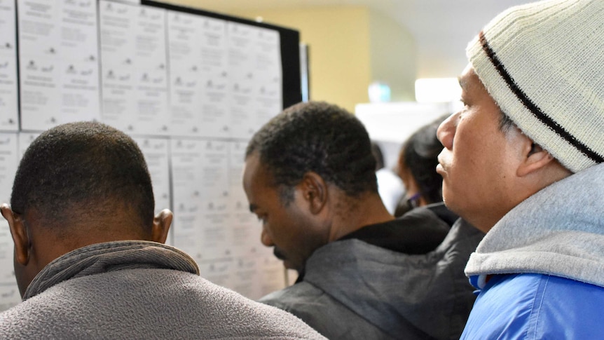 A group of men crowd around a board posted with job ads at the Sunshine Jobs Fair.